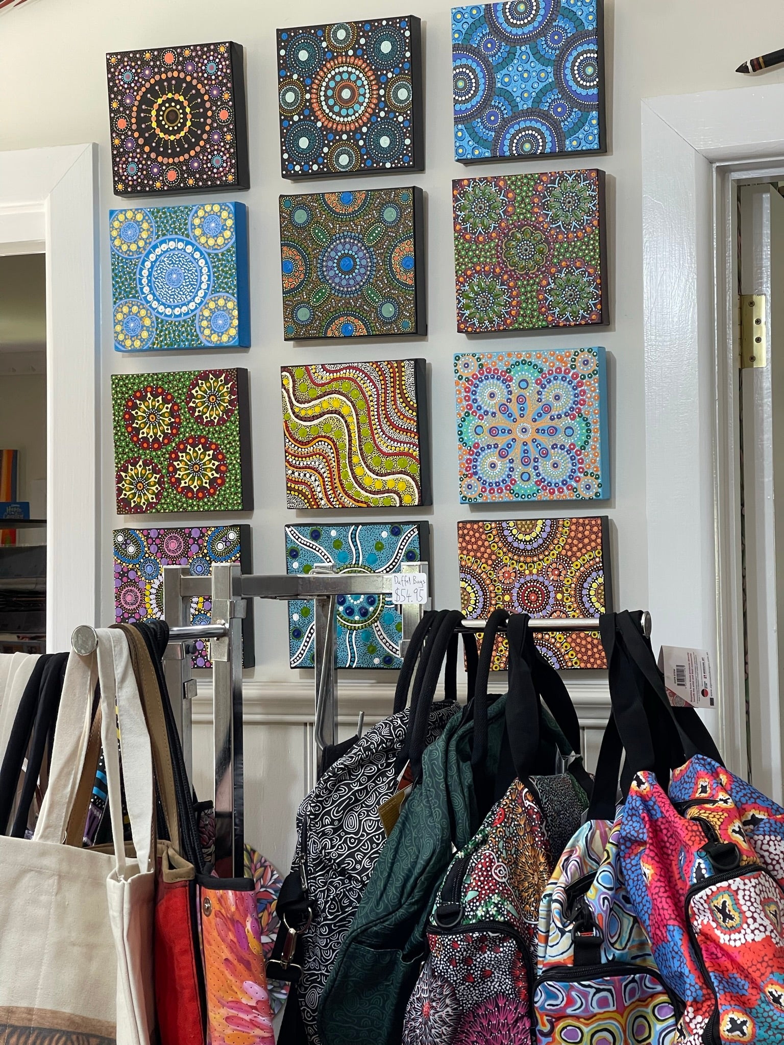 Image of small pieces of art hanging on a wall with a stand of bags in front. Picture from Raintree Art, Aboriginal Art Ballarat