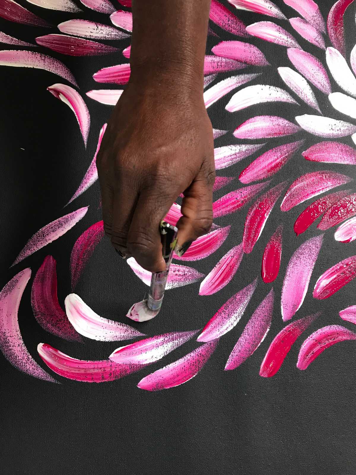 Woman's hand holding brush, placing white and pink paint on a black canvas. Image taken at Raintree Art, Aboriginal Art Ballarat